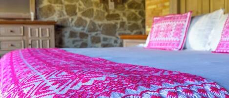 Quarto com parede de pedra, cabeceira de madeira e cama com manta estampada em rosa e branco.