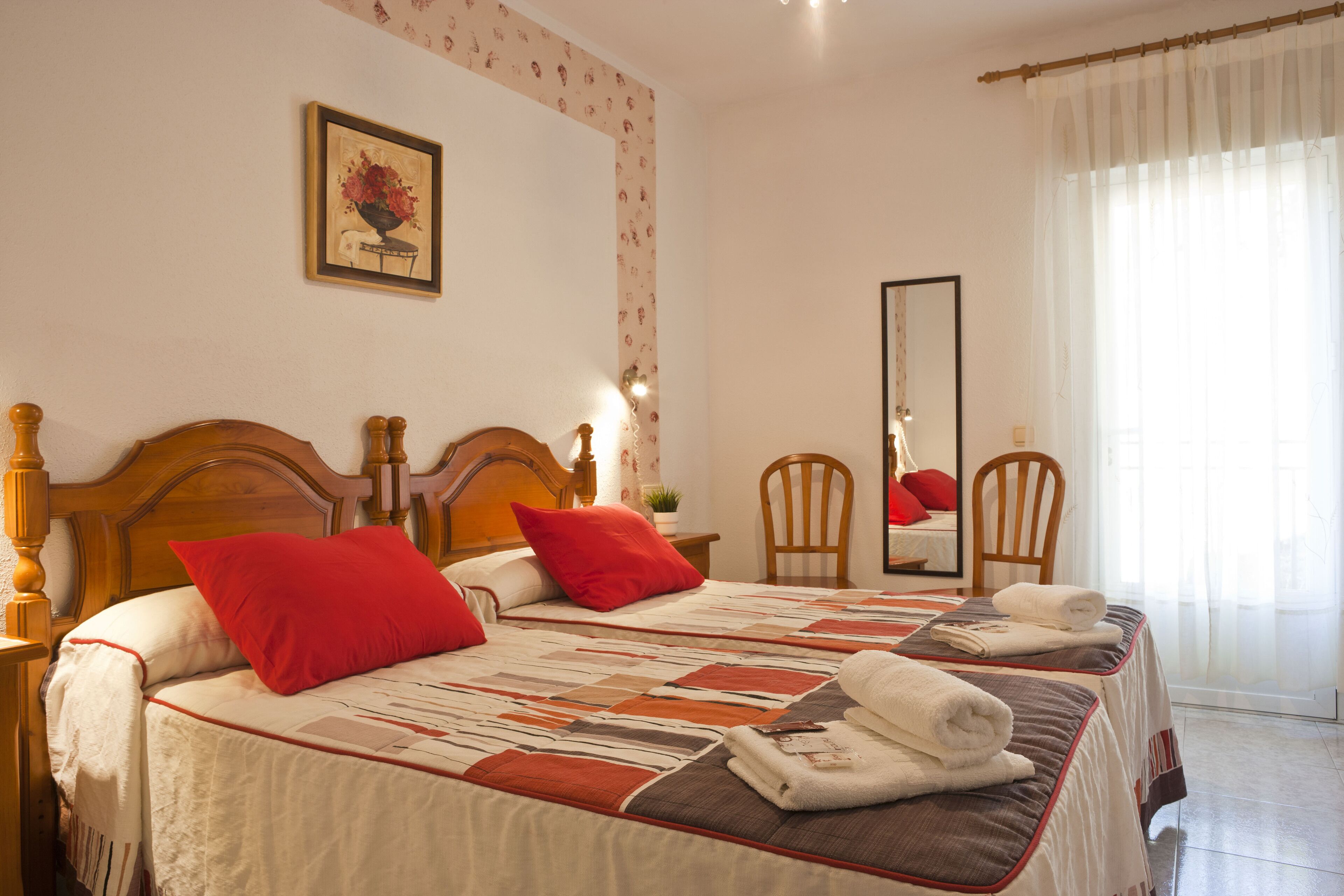 Un lit double bien fait, avec une literie rouge et blanche, une tête de lit en bois et une courtepointe. On y trouve une table de chevet en bois avec une lampe, une chaise et un miroir accroché au mur.
