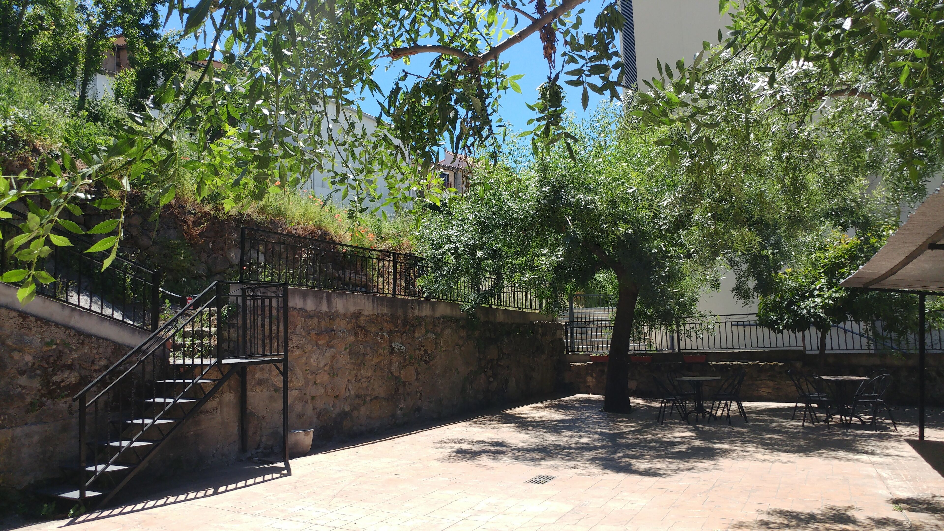 Un patio con muro de piedra, escalera de metal, zona de estar al aire libre y árboles que dan sombra.