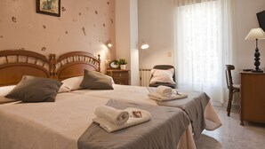 A hotel room with two beds, a wooden headboard, a bedside table with a lamp, a chair, and a desk with a lamp.