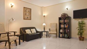 A living room with a black sofa, two armchairs, a small table, a floor lamp, a bookshelf, a potted plant, and a flat-screen TV mounted on the wall.