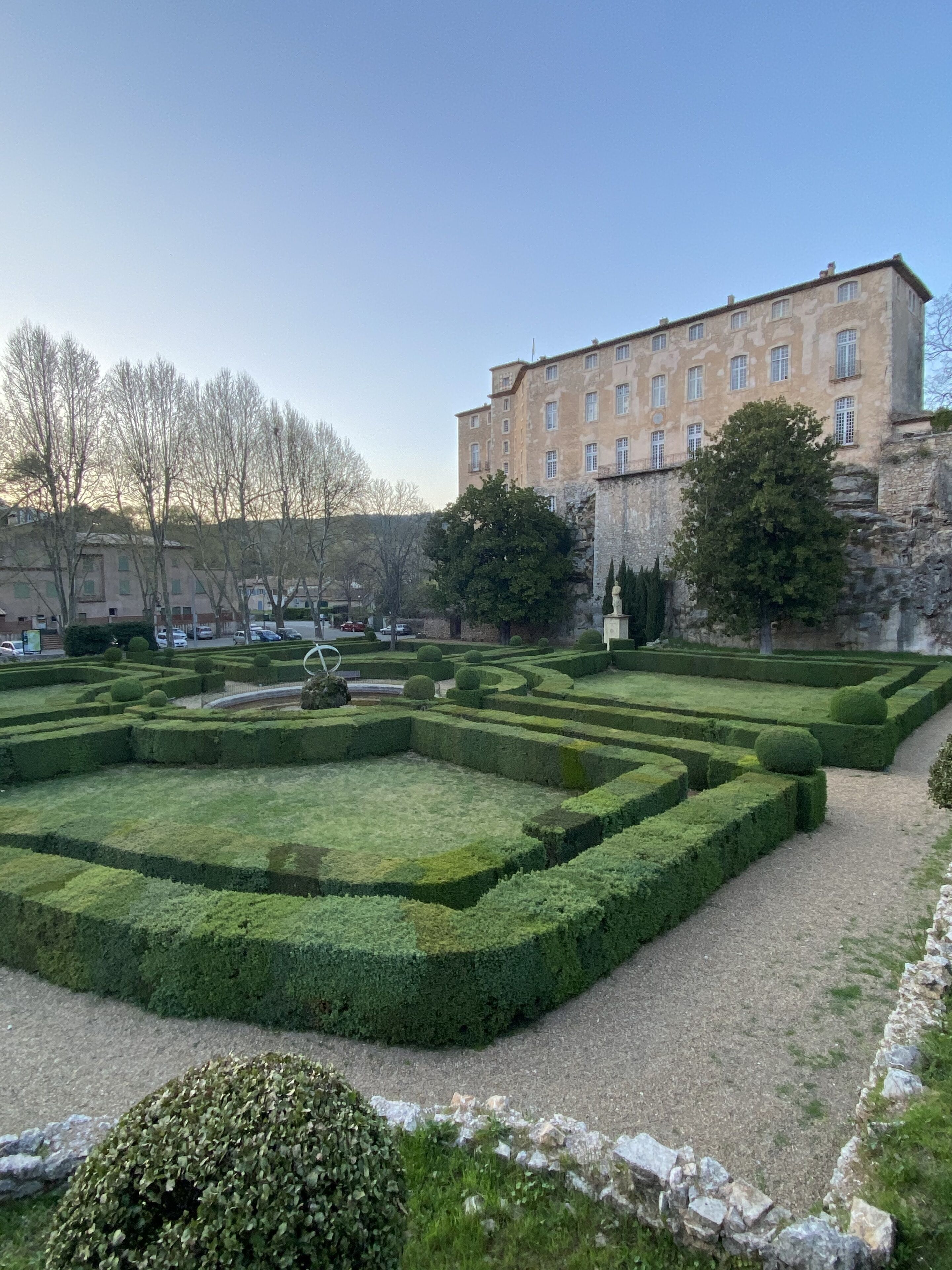Un jardín formal con setos geométricos, una escultura central y un edificio histórico al fondo.