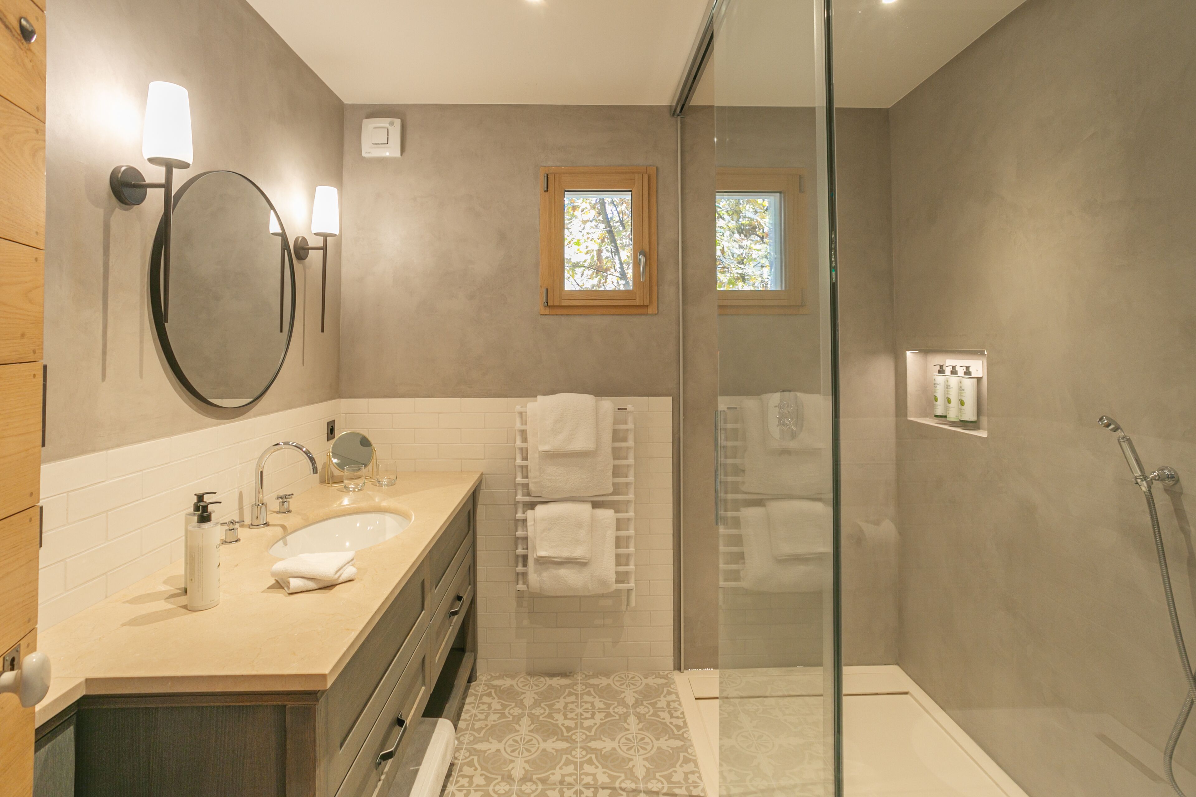 A modern bathroom with a large mirror, two sinks, a glass-enclosed shower, and a small window.