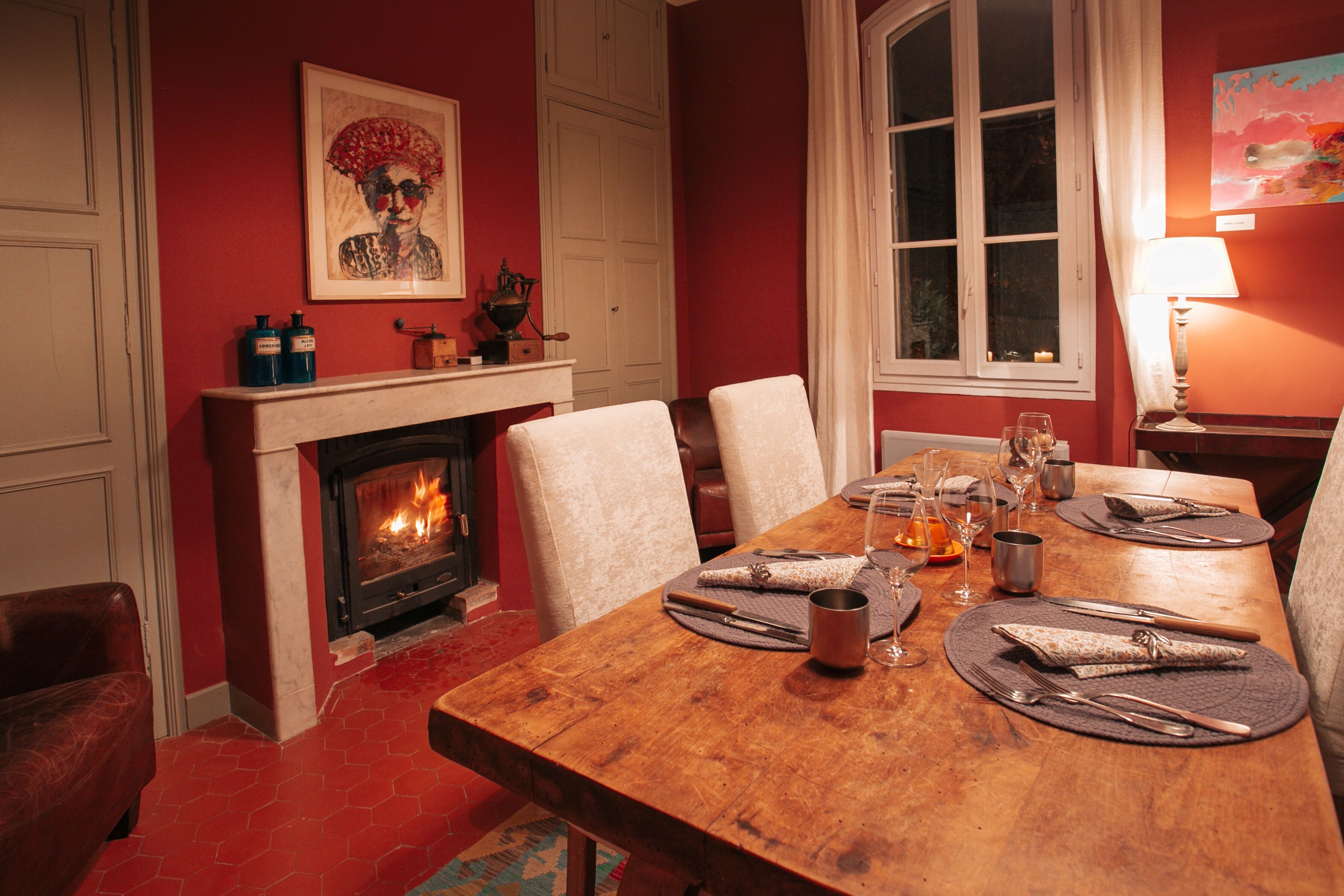 Une salle à manger avec une table en bois dressée pour quatre personnes, une cheminée et une grande fenêtre avec des rideaux.