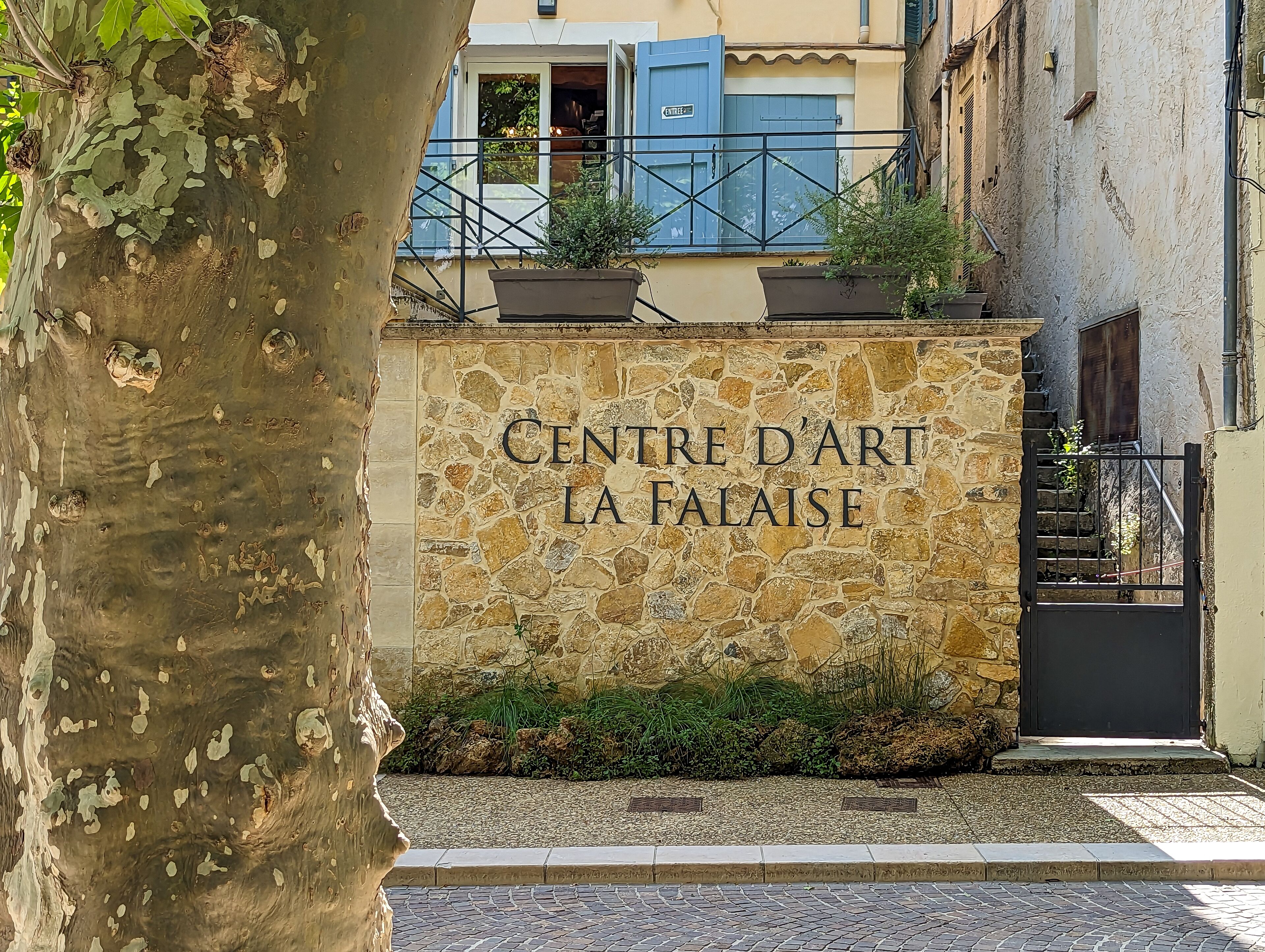 A stone wall with the inscription "Centre d'Art La Falaise" and a gate leading to a building with a blue door.