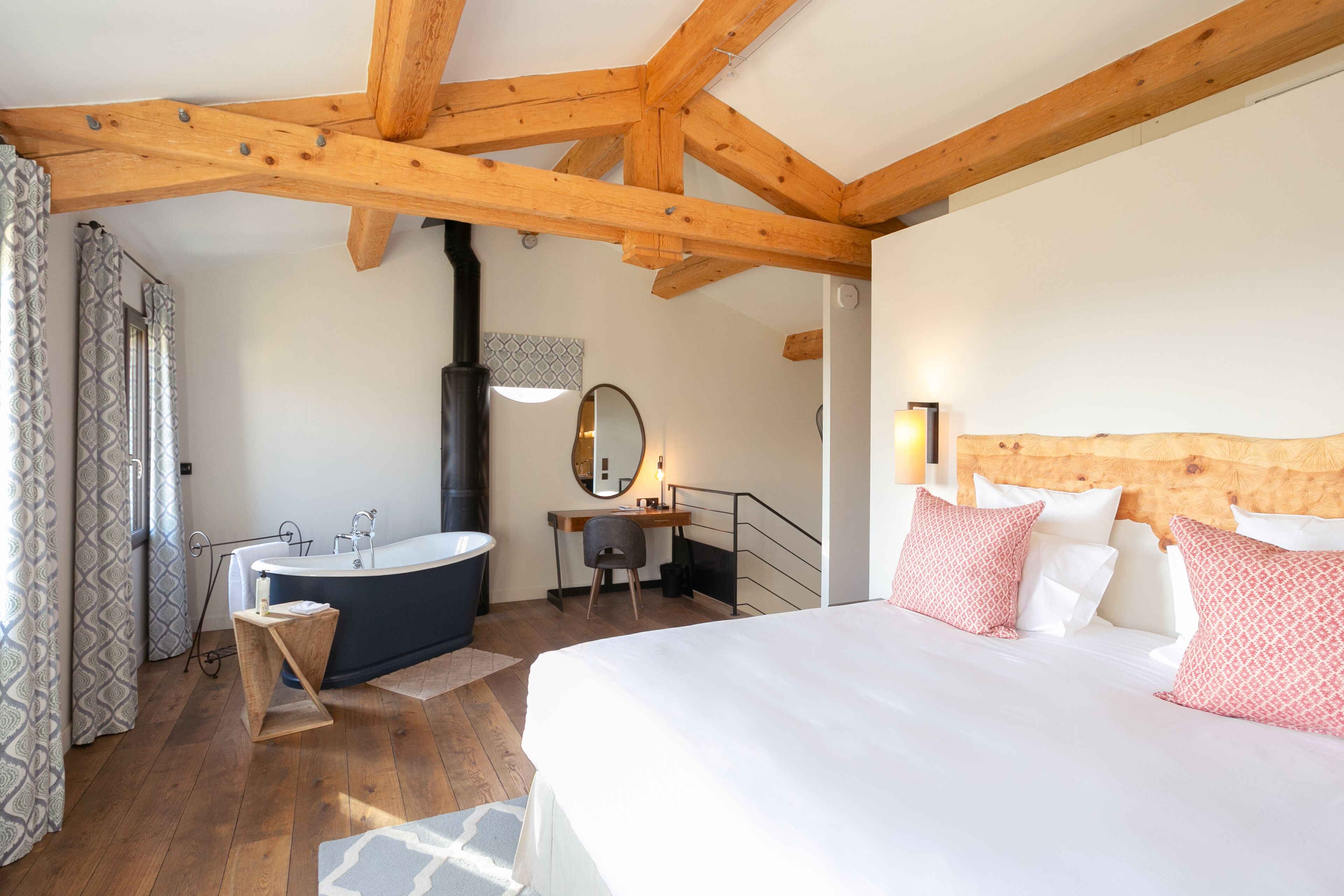 A bedroom with a large bed, a wooden ceiling, a bathroom with a bathtub, and a desk.