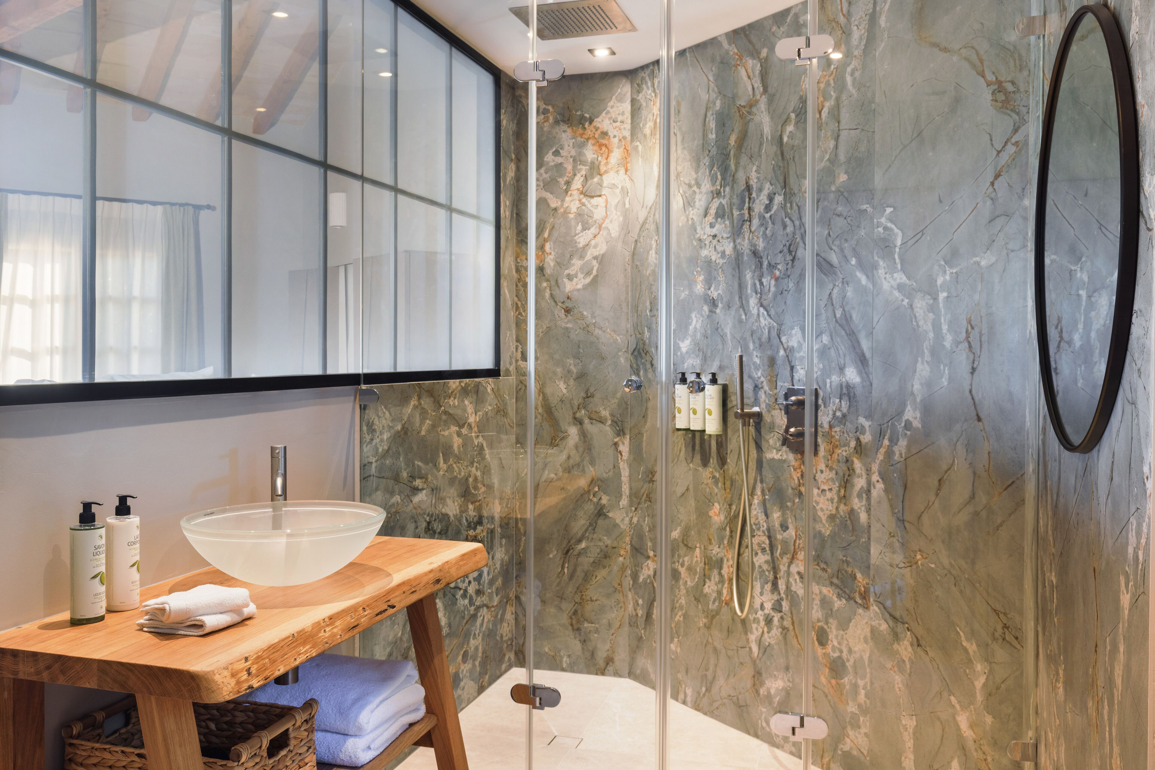 Un baño con ducha de vidrio, tocador de madera con un lavabo grande y pared de mármol.
