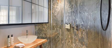 Un baño con ducha de vidrio, tocador de madera con un lavabo grande y pared de mármol.