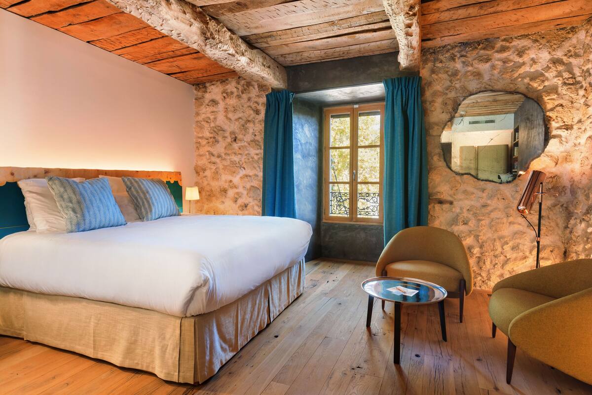 A bedroom with a bed, two chairs, a small table, a window with curtains, and a stone wall.