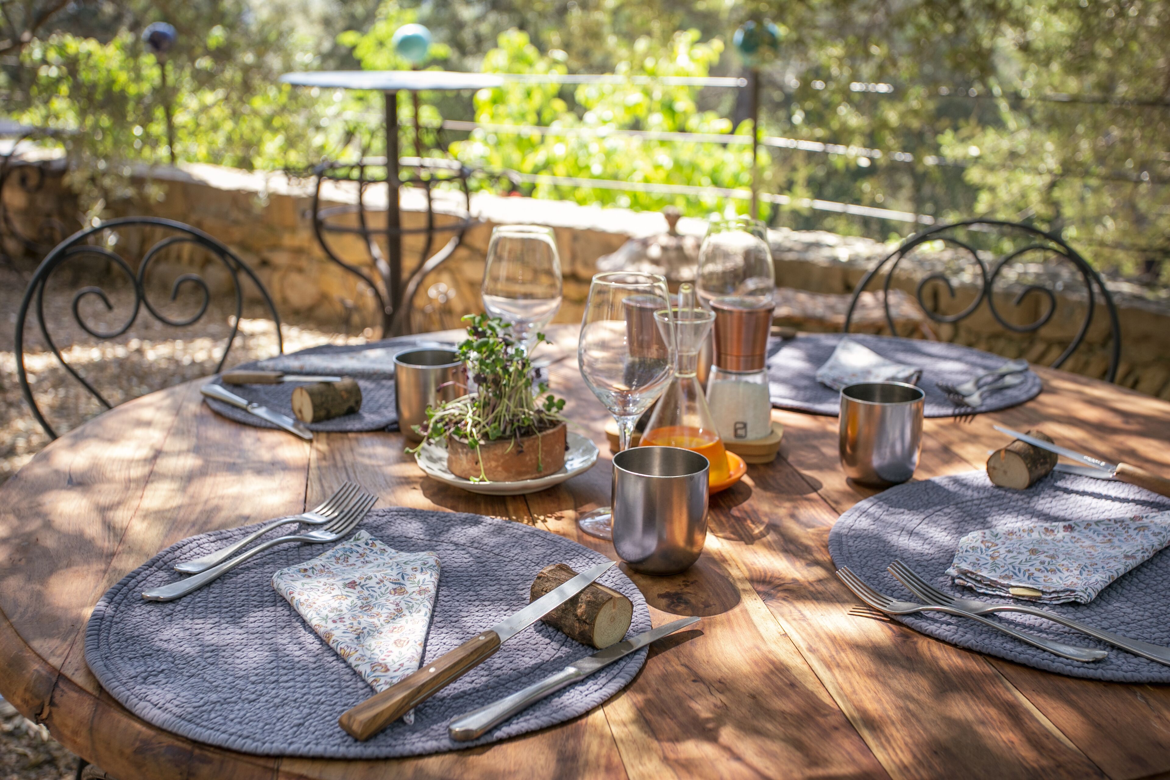 A round wooden table set outdoors with metal cutlery, a small plant, and a glass pitcher.