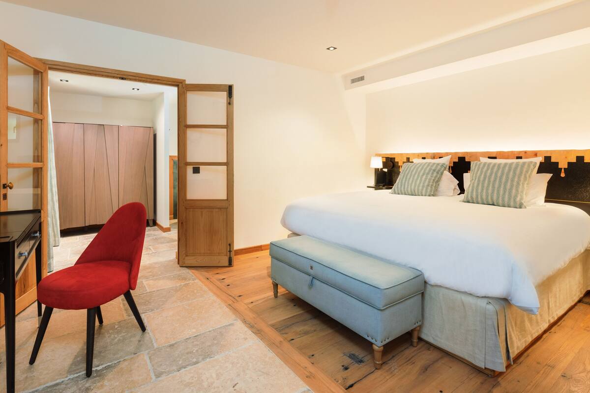 A bedroom with a bed, a red chair, a wooden wardrobe, and a stone floor.