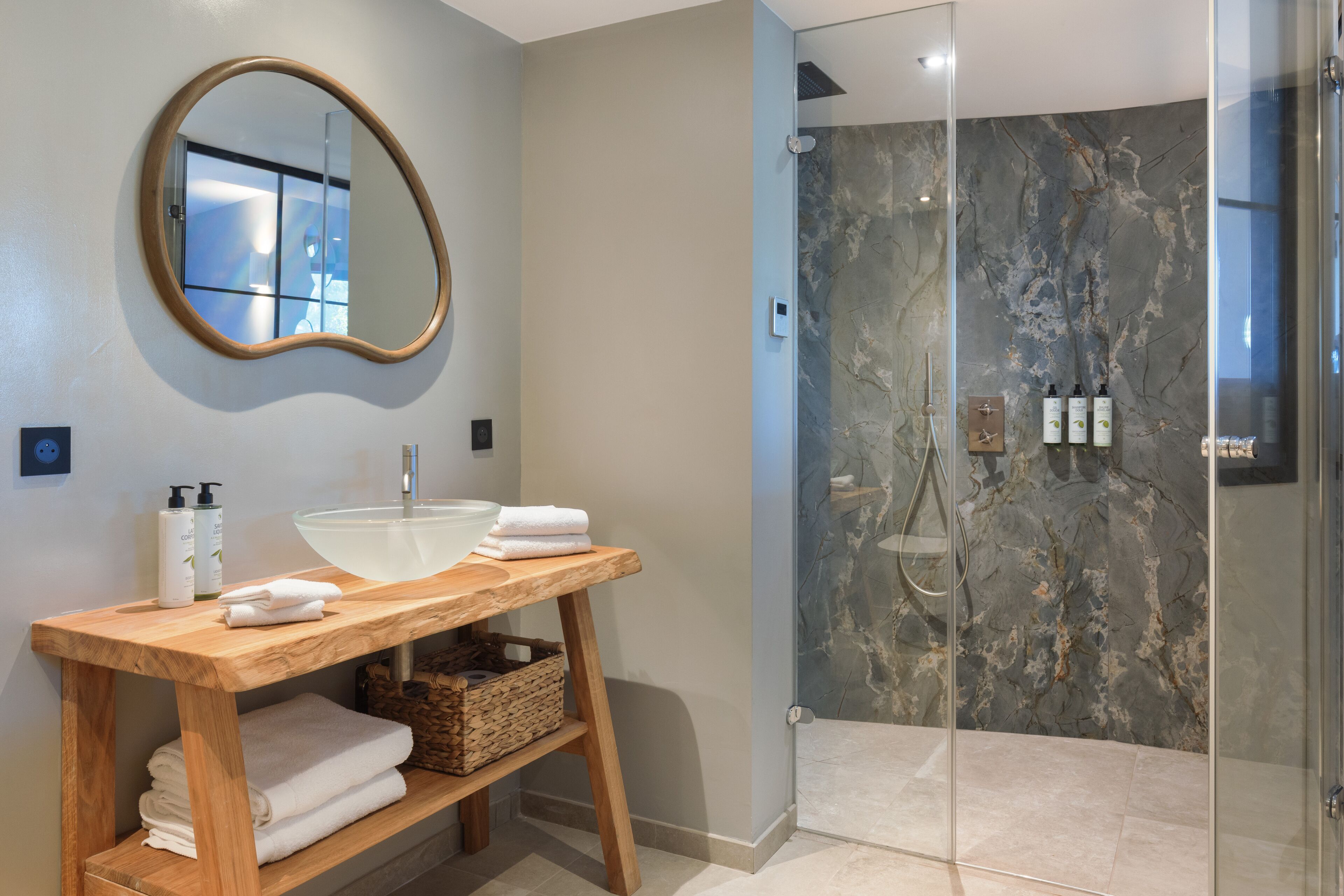 A modern bathroom with a wooden vanity, a large mirror, a glass-enclosed shower, and marble walls.