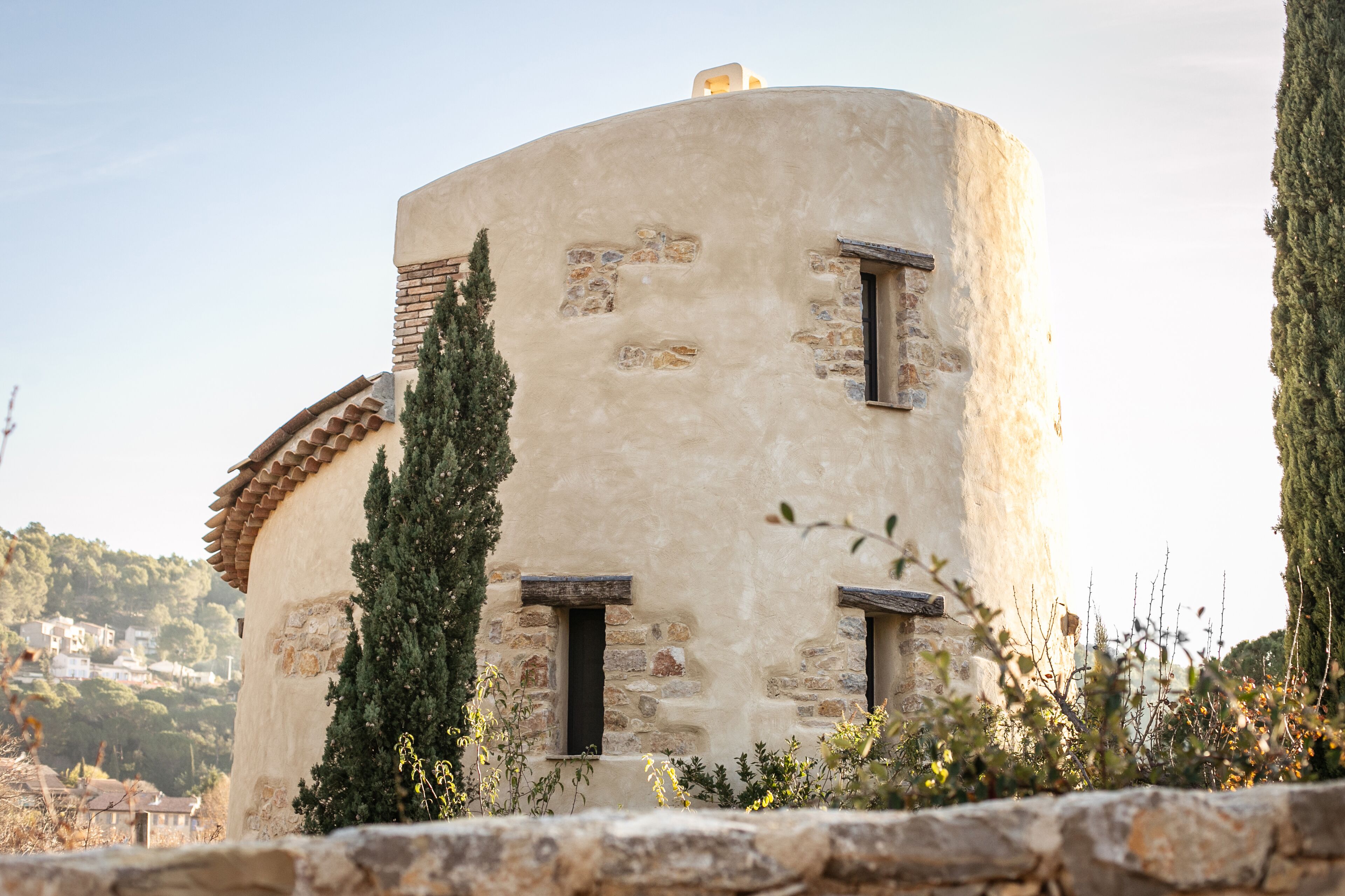 Un edificio de piedra cilíndrico con dos ventanas y una pequeña puerta, rodeado de vegetación y un muro de piedra.