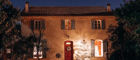 Un edificio de dos pisos con una puerta roja, varias ventanas con contraventanas y un tejado de tejas.