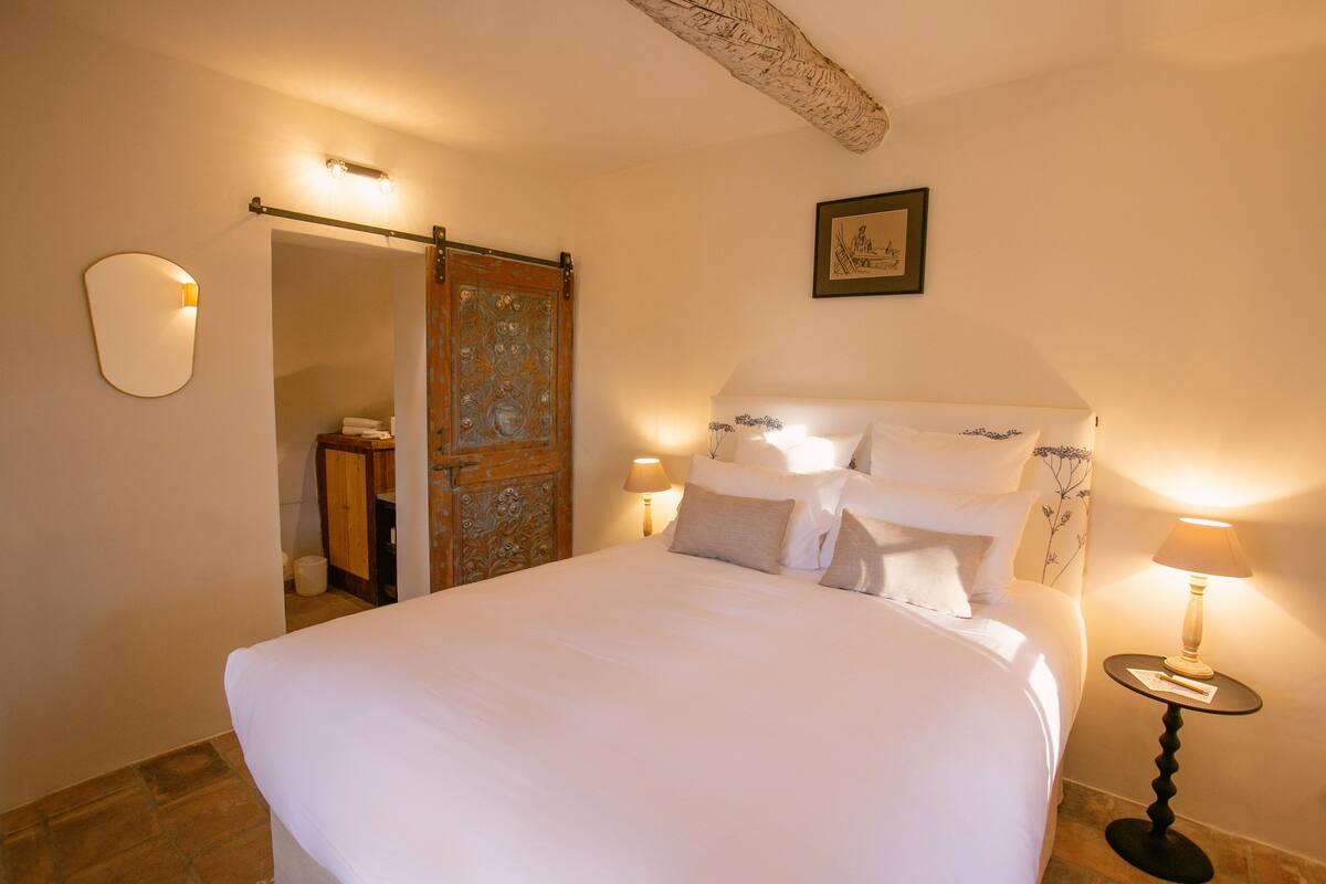 A neatly made bed with white linens, a wooden door with intricate carvings, a framed picture on the wall, and two bedside lamps.