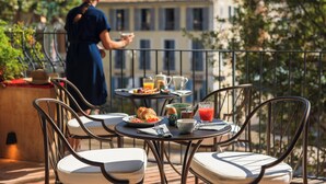 Ein Balkon mit Sitzgelegenheit im Freien, einem Tisch mit Frühstücksangeboten und Blick auf Gebäude im Hintergrund.