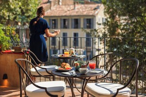 Un balcón con mobiliario exterior, una mesa con alimentos para el desayuno y vistas a edificios al fondo.