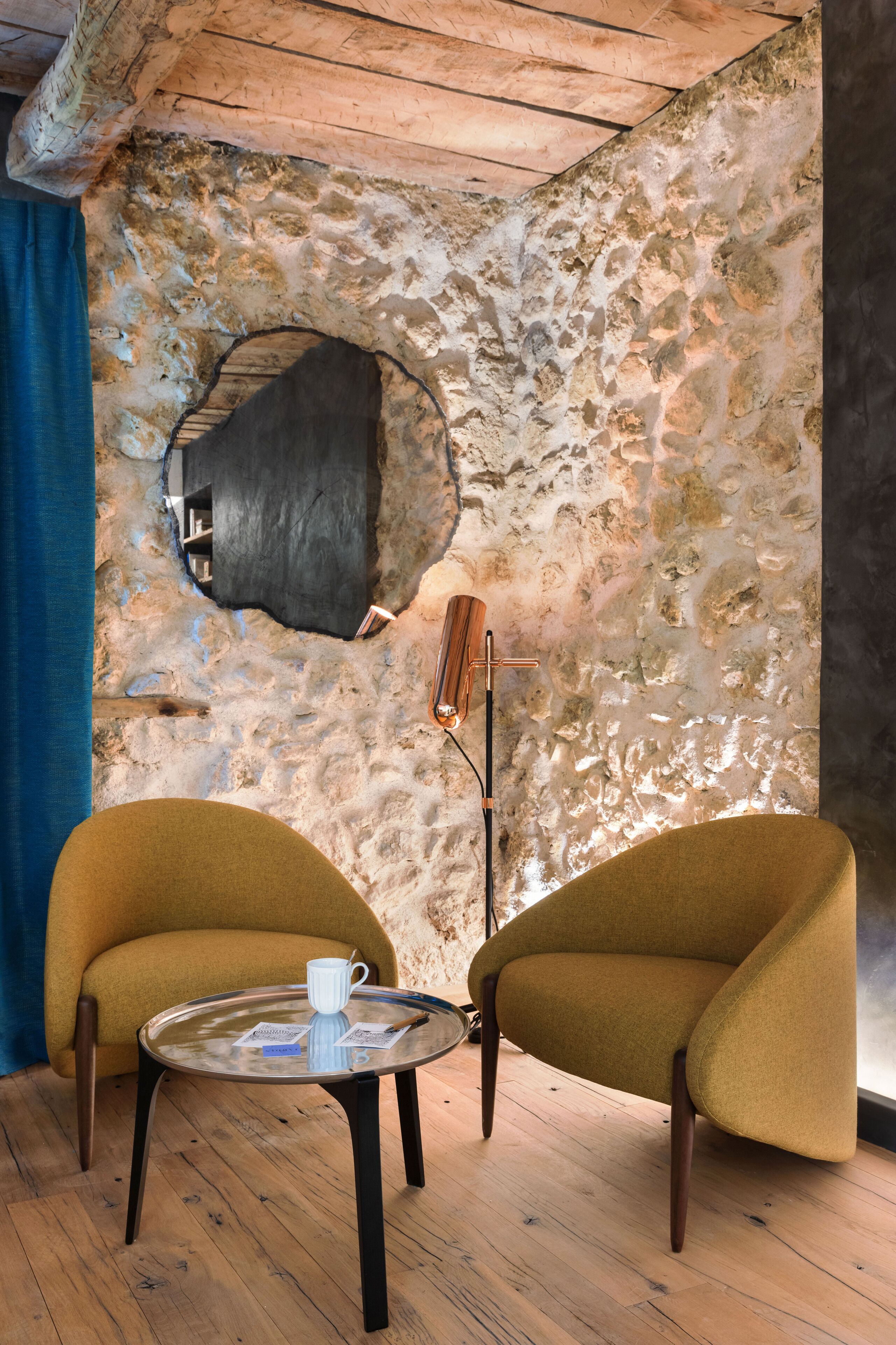 Una habitación con pared de piedra, un espejo redondo, una mesa con una taza y dos sillones.