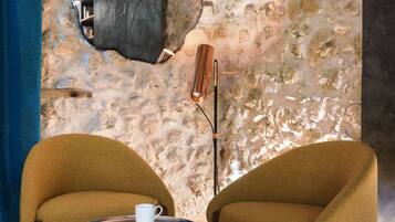 Una habitación con pared de piedra, un espejo redondo, una mesa con una taza y dos sillones.