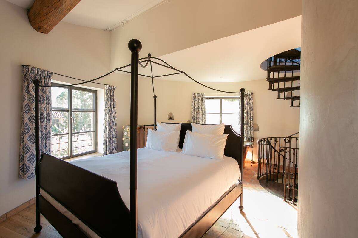 A four-poster bed with white linens, a wooden headboard, and a view of a spiral staircase.