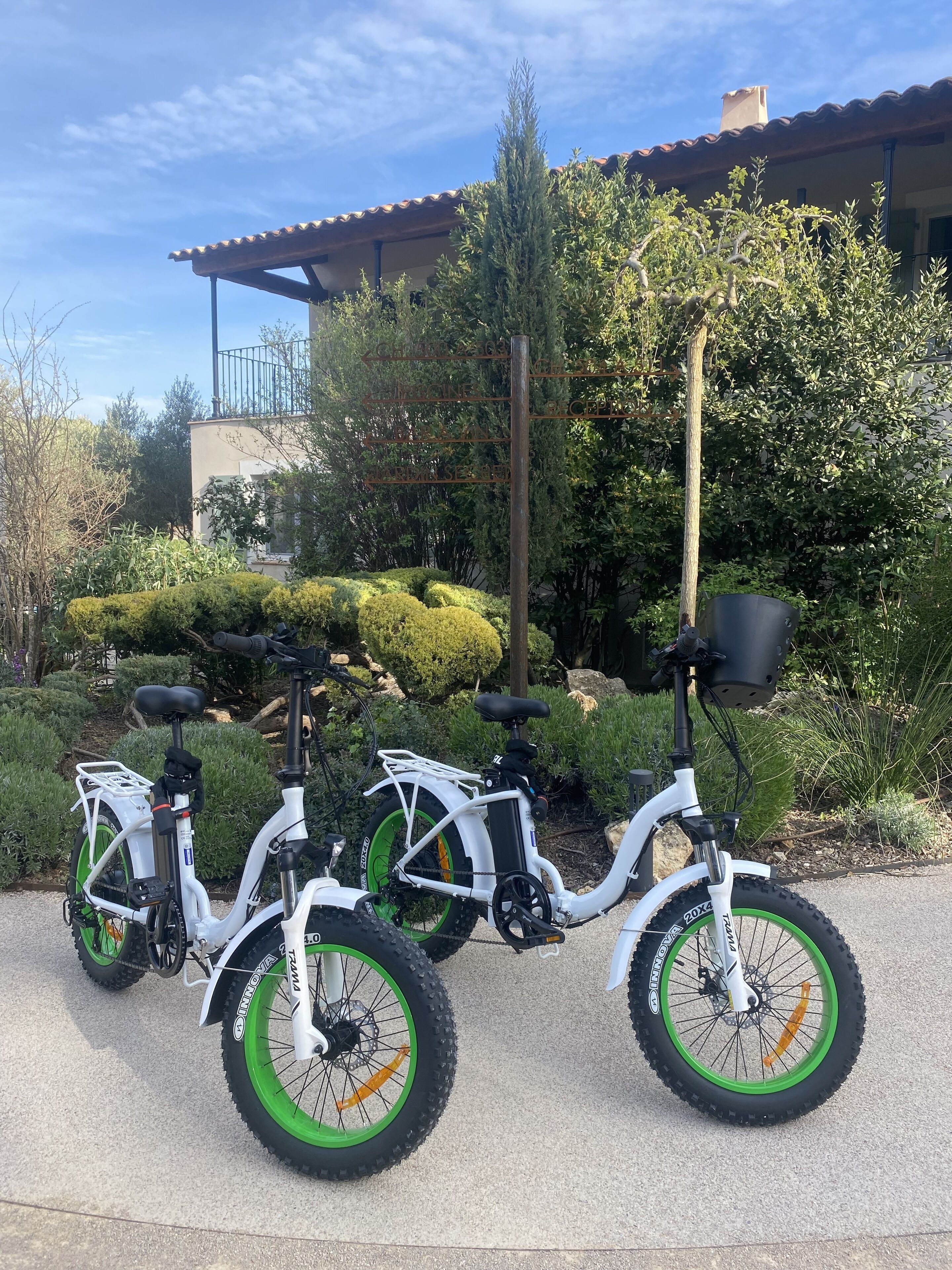 Drei weiße E-Bikes stehen in einer Auffahrt mit grünen Akzenten vor einem Gebäude mit einem Ziegeldach und einem Metallzaun.
