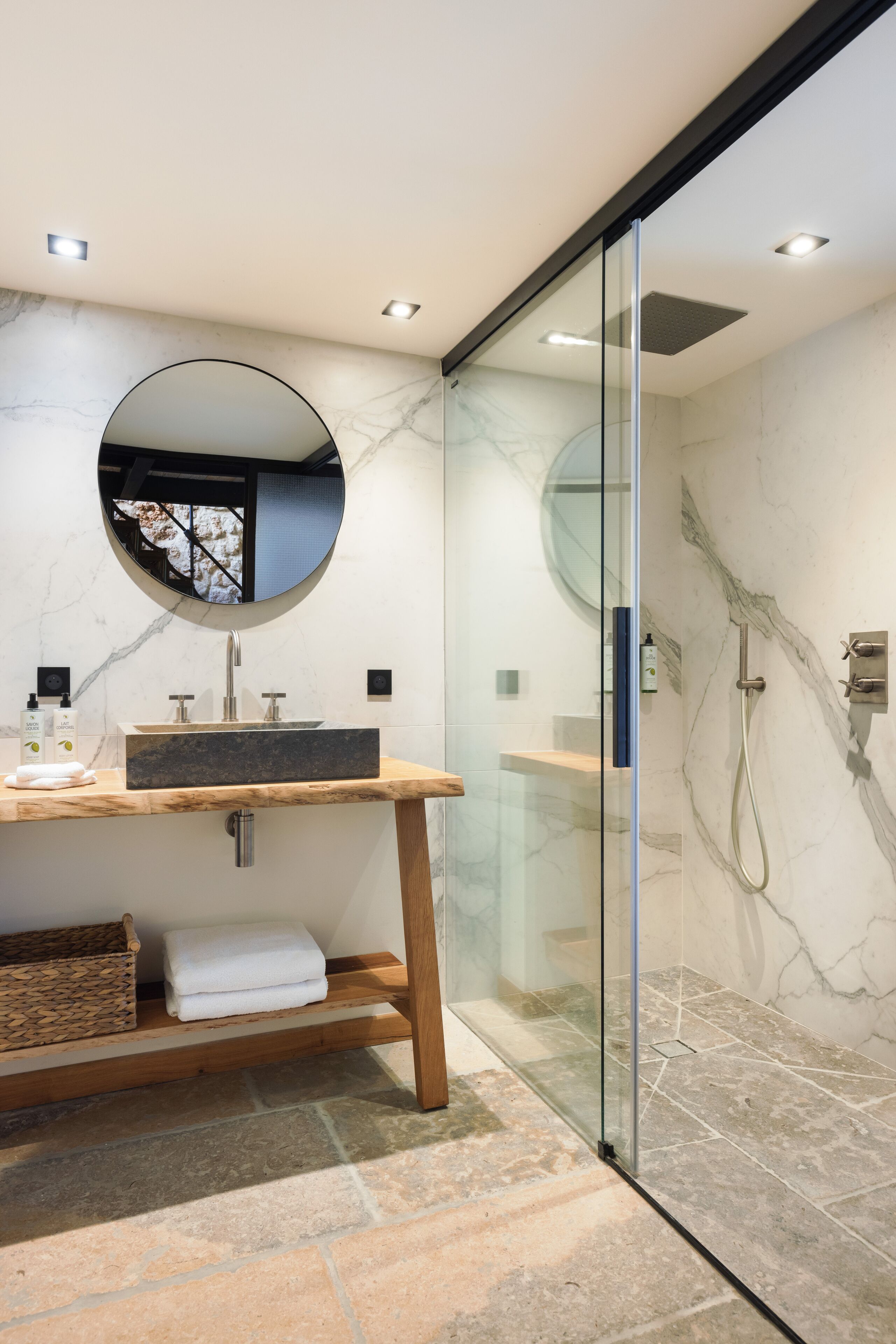 Une salle de bain moderne avec un lavabo en marbre, un miroir rond et une douche séparée par des parois en verre.