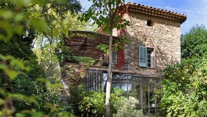 Ein rustikales Steinhaus mit einem hölzernen Balkon und einer Glaseinfassung, umgeben von üppiger Vegetation.