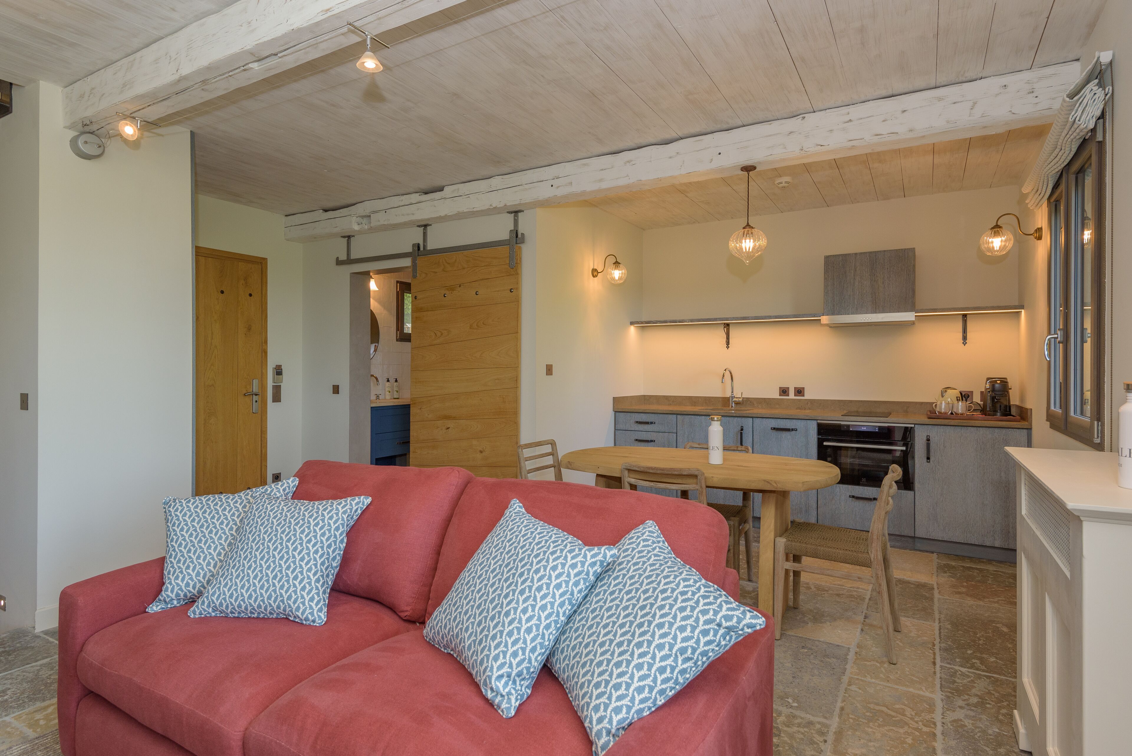 A cozy living area with a red sofa and patterned cushions, a wooden dining table, and a kitchen with modern appliances and pendant lighting.