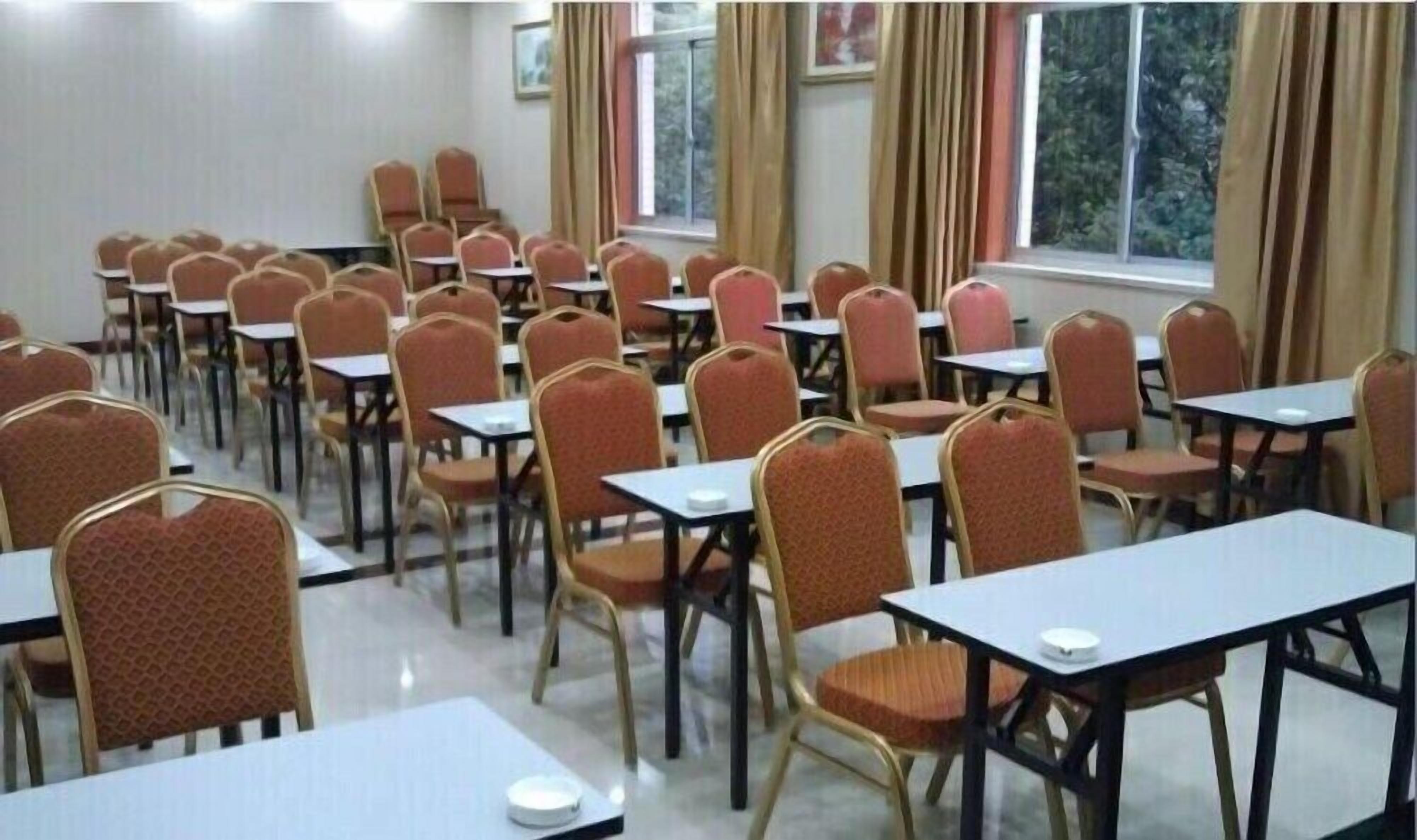 A conference room with rows of tables and chairs, large windows with curtains, and a tiled floor.