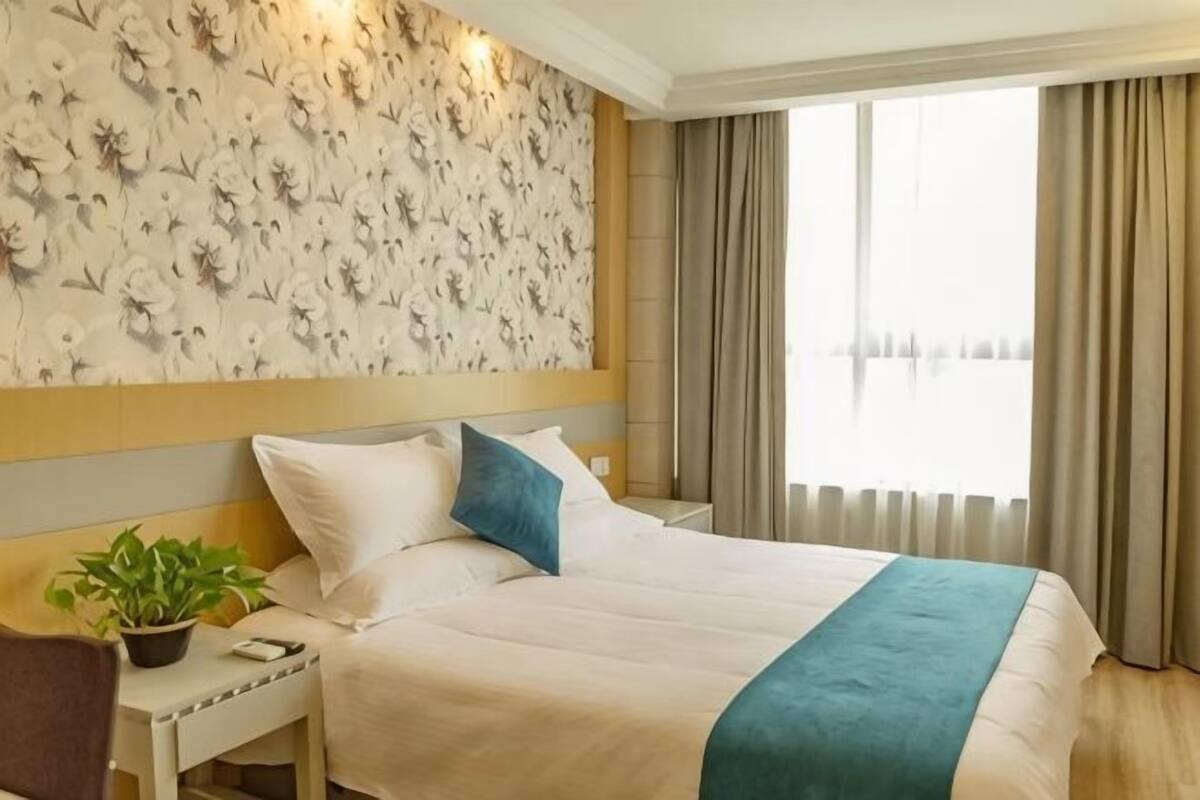 A hotel room with a large bed, a nightstand, a potted plant, and a window with curtains.
