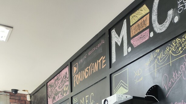 A person seated at a wooden desk in a room with a blackboard wall covered in various writings and drawings. - MEC Office Hostel (Niteroi)
