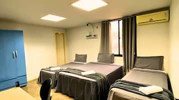 Quarto de hotel com duas camas, uma mesa de trabalho e uma cadeira. Há uma janela com cortinas, uma lâmpada no teto e um aparelho de ar condicionado.