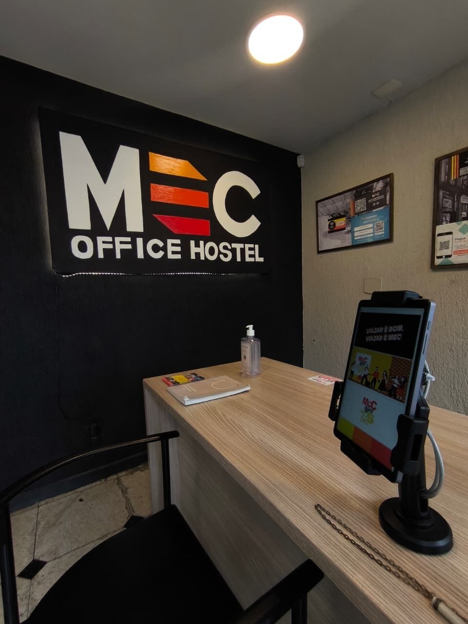 Zona de recepción de un hostal tipo oficina, con un letrero que dice 'MEC OFFICE HOSTEL', un escritorio de madera, una botella de gel antibacterial y un quiosco informativo digital.