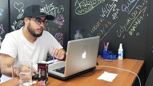 A man at a wooden desk with a laptop, a glass of water, and a YoPro energy drink box. The wall behind him is covered with colorful chalk writings and drawings. - MEC Office Hostel (Niteroi)
