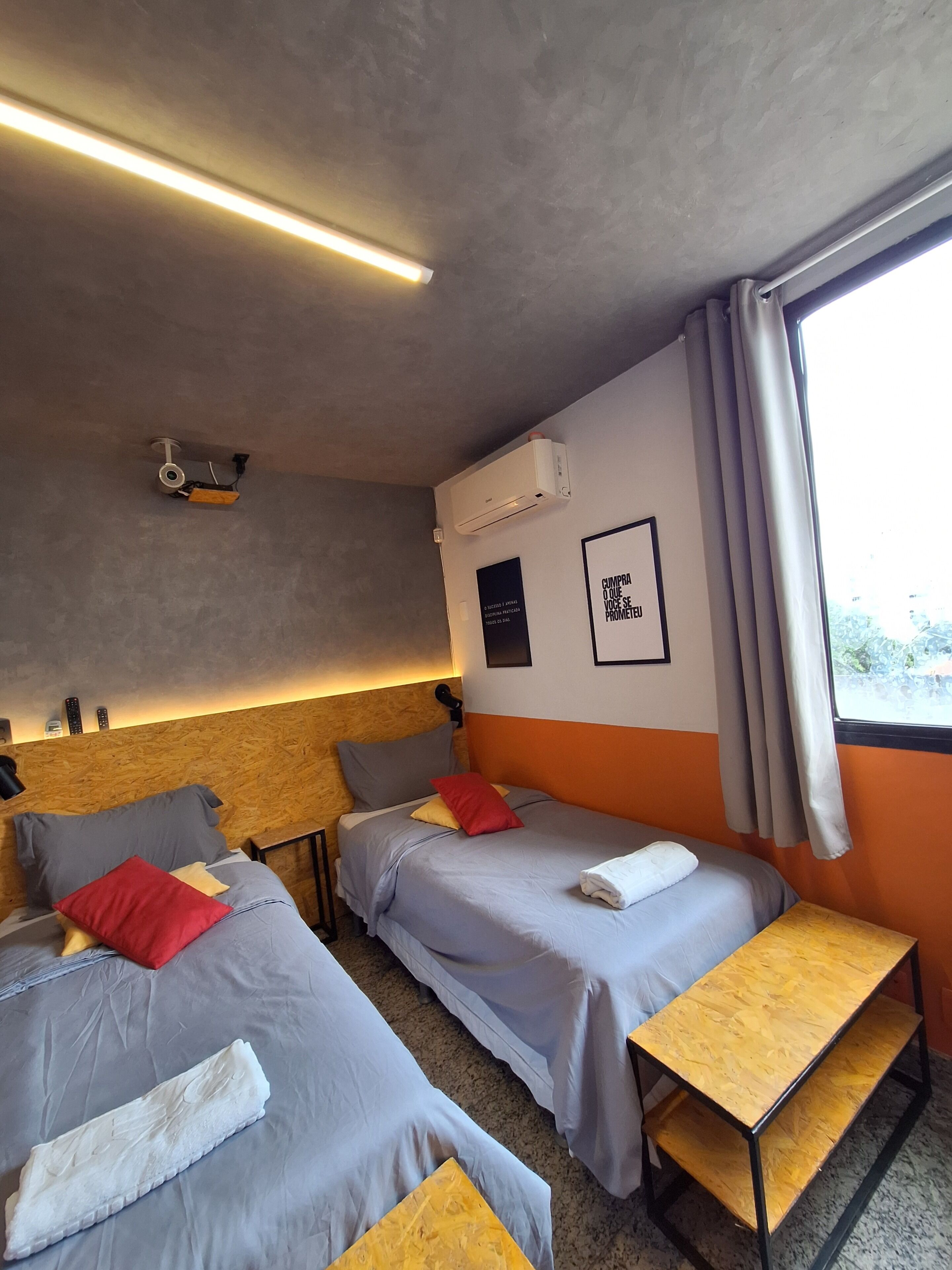 Habitación de hotel con dos camas, cabecero de madera, aire acondicionado, una cita enmarcada en la pared y una ventana con cortinas.