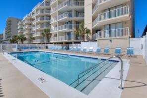 Una piscina con sillones de descanso y una cerca blanca, rodeada por un edificio de varios pisos con balcones.