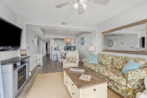 A spacious living room with a sofa, coffee table, and a television. There is a kitchen area in the background with a dining table and chairs.