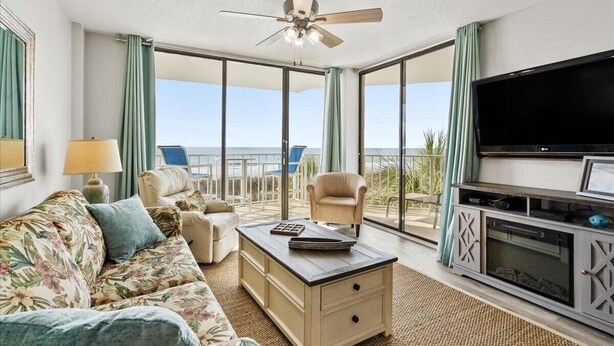 A spacious living room with a ceiling fan, a flat-screen TV, a sofa, a coffee table, and a balcony with a view of the beach. - Oceanfront 2Bed 2Bath Condo with Large Balcony & Amazing views  (North Myrtle Beach)