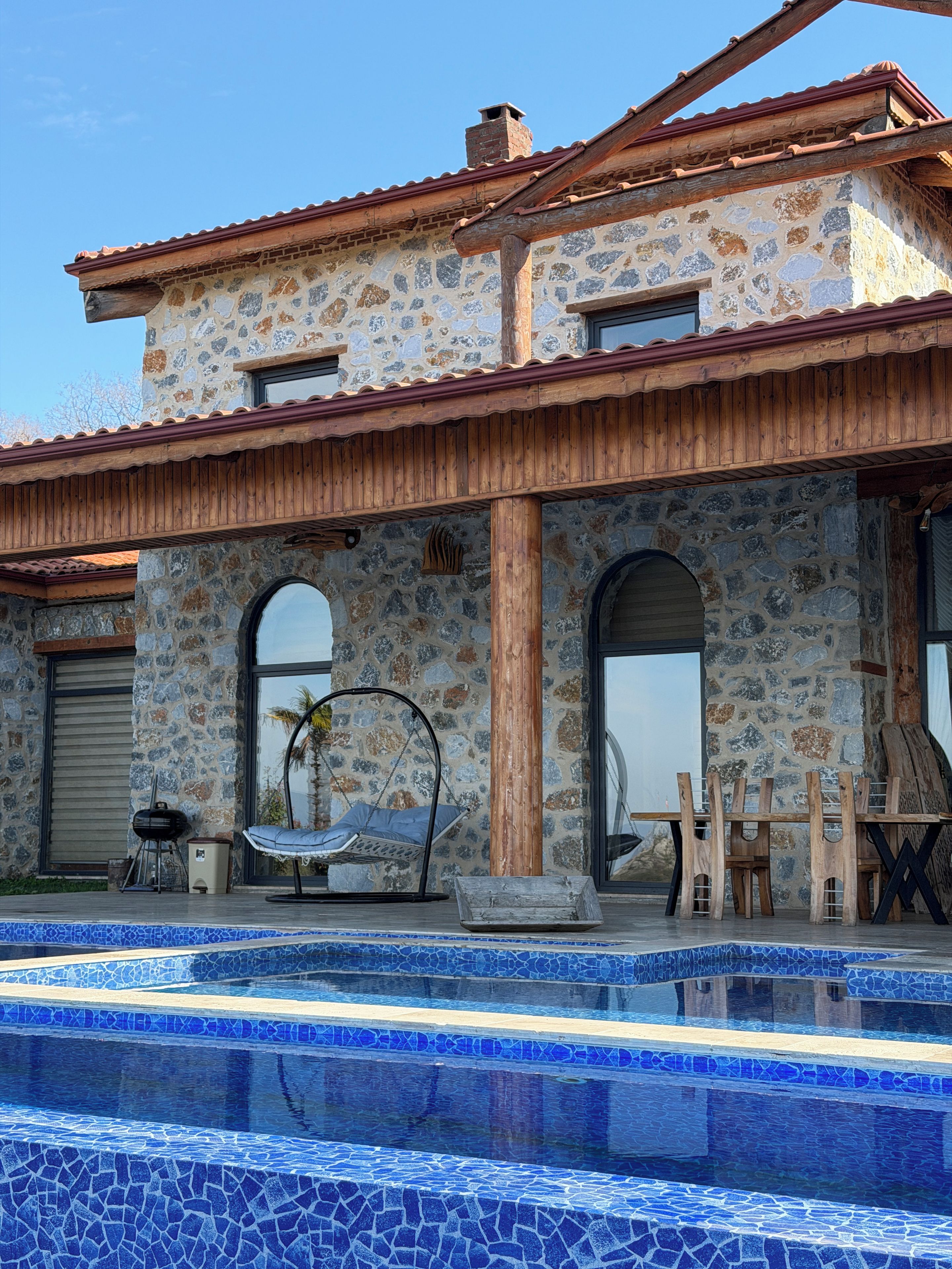 Una casa de piedra de dos pisos con piscina, terraza de madera y mobiliario exterior.