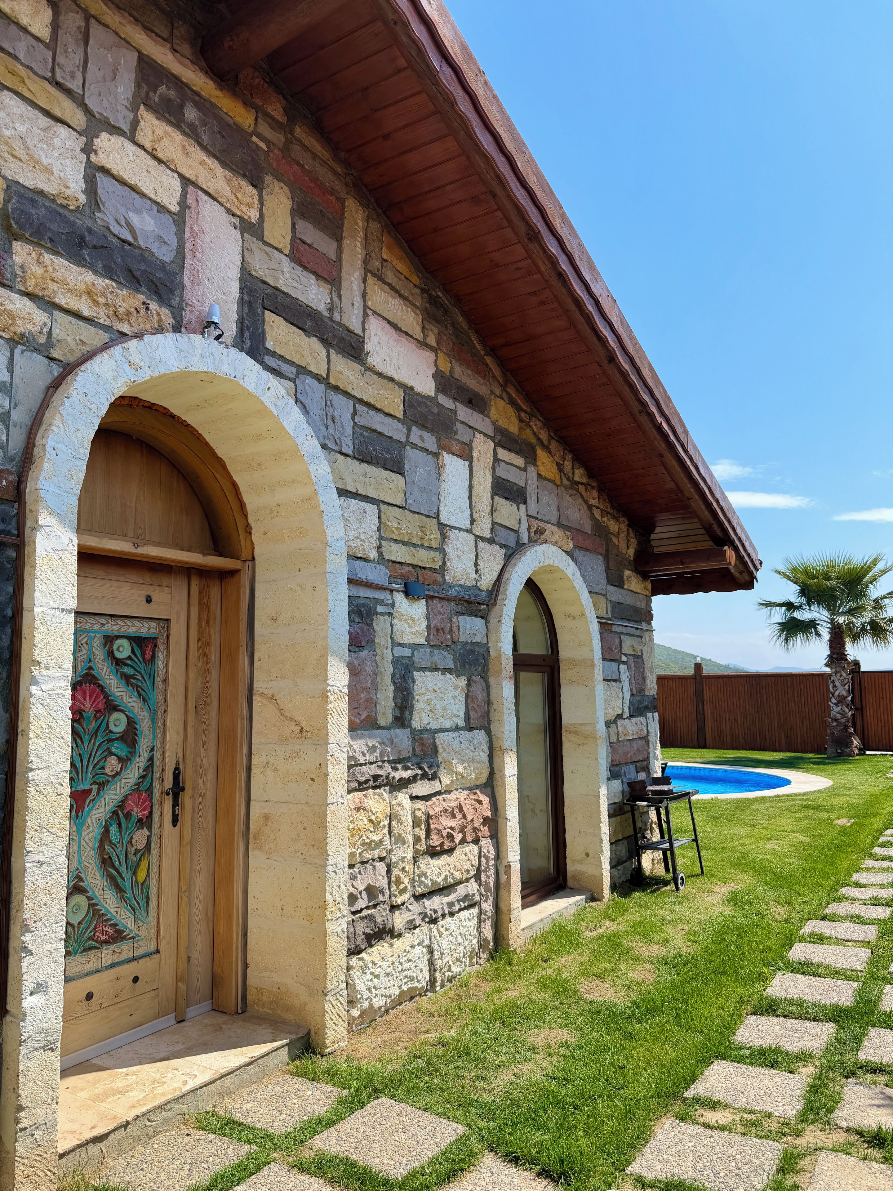 Um edifício de pedra com porta de madeira e janelas em arco, piscina e gramado bem cuidado.