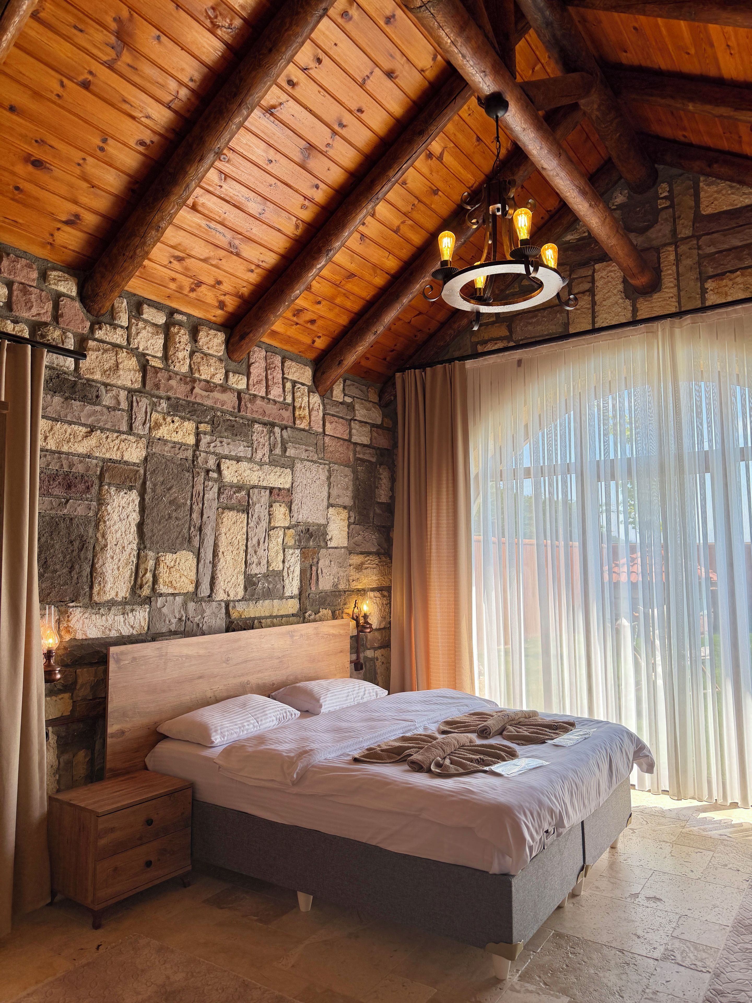 Un dormitorio con pared de piedra, vigas de madera en el techo, una cama con almohadas y una colcha, una mesita de noche y una lámpara colgante.