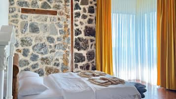 Quarto com parede de pedra, vigas de madeira no teto, cama com roupa branca e janela com cortinas amarelas.