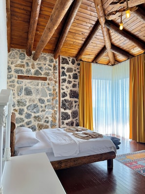 Un dormitorio con pared de piedra, vigas de madera en el techo, cama con ropa blanca y una ventana con cortinas amarillas.