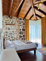 Quarto com parede de pedra, vigas de madeira no teto, cama com roupa branca e janela com cortinas amarelas.