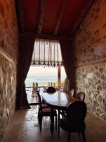 Una habitación con paredes de piedra, una mesa y sillas de madera, vista al mar a través de una ventana con cortinas, y piso de piedra.