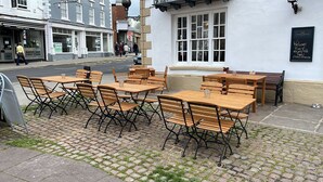 Outdoor wooden tables and chairs on a cobblestone pavement, with a white building in the background. - Punch House (Monmouth)