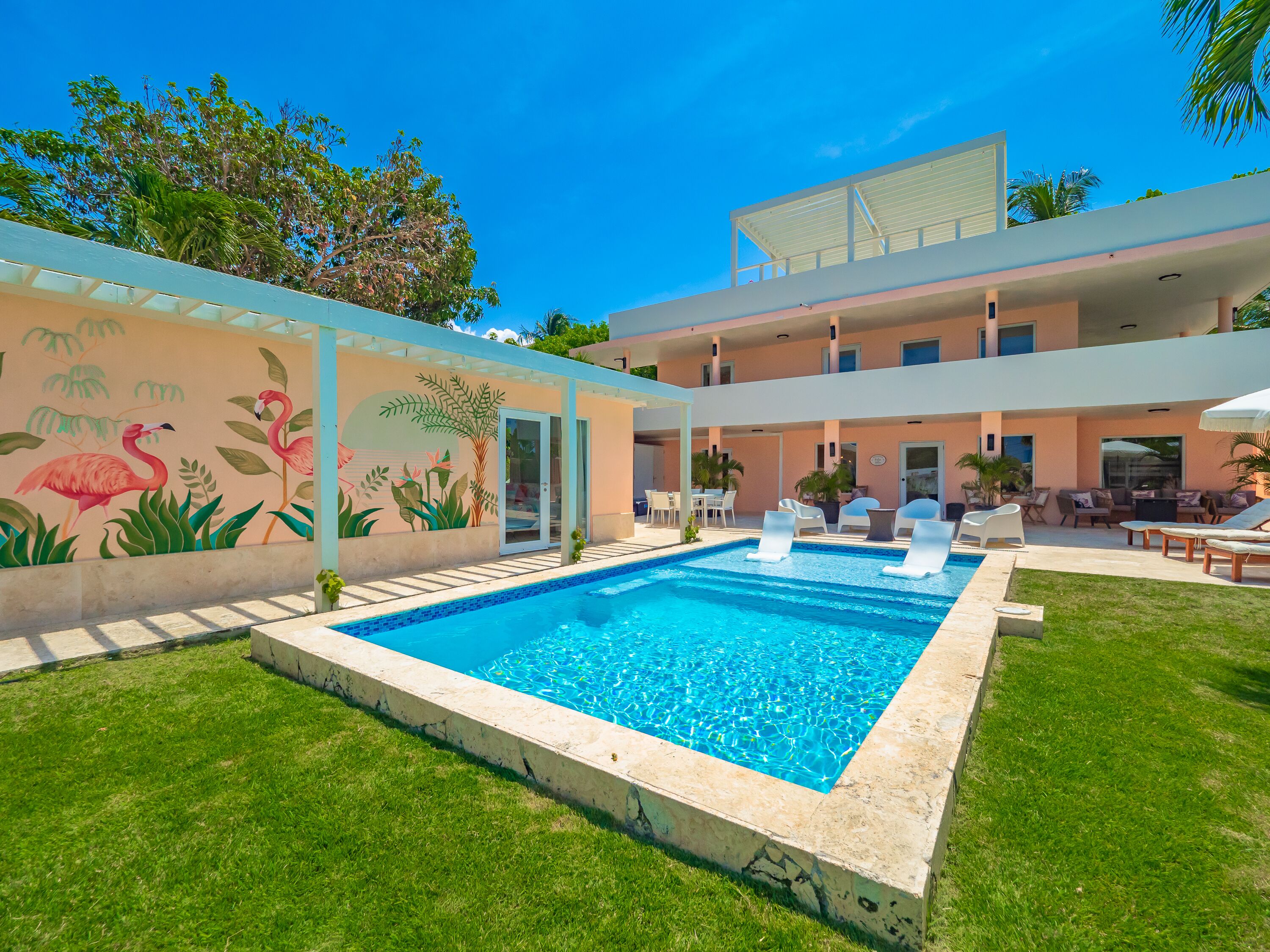 Ein moderner Poolbereich mit einem klaren blauen Pool, Sitzmöglichkeiten im Freien und einem Wandbild von Flamingos und Pflanzen.