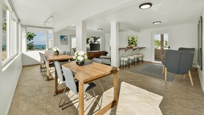 A spacious living area with a wooden dining table, grey chairs, and a grey sofa. There is a kitchen area with a counter and stools, and a large window offering a view of the outdoors. - Palm Villa- Rooftop, Pool and Direct Beach Access (Río Grande)