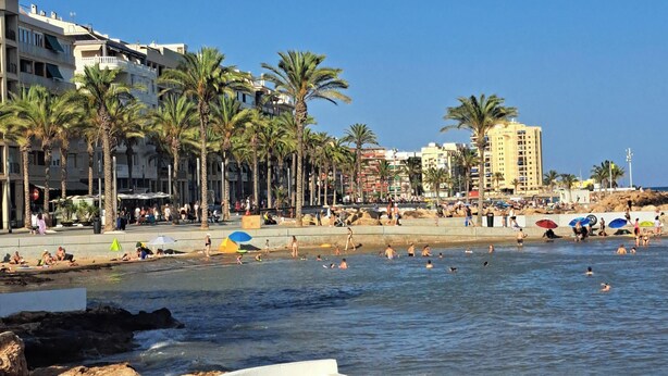 Een strand met palmbomen, mensen die zwemmen en gebouwen op de achtergrond.
