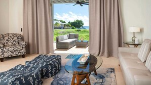 A room with a view of a grassy area and a body of water, a sofa, a patterned armchair, a glass-top table, and a lamp. - Pirates Villa- Beach view with kayaks for Groups (Cruz Bay)